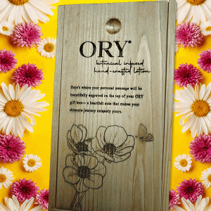 ORY wooden gift box with engraved logo and floral illustration, displayed on a vibrant yellow background framed by daisies and pink flowers, highlighting personalized botanical skincare gifting.