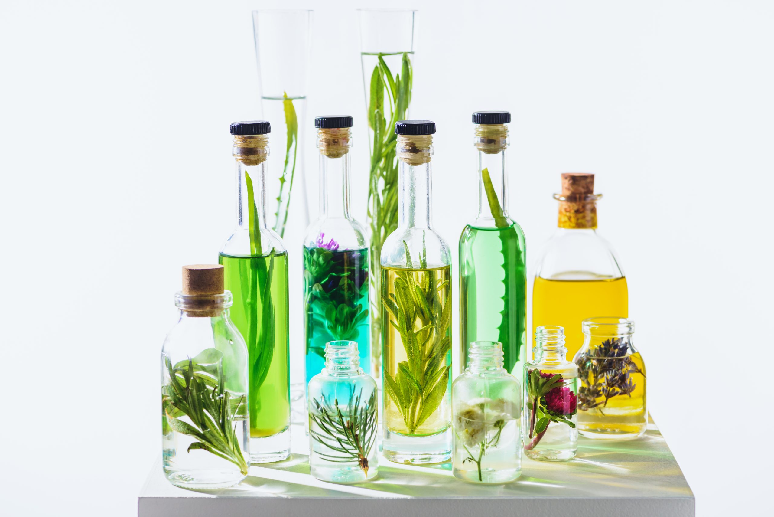 Collection of glass bottles filled with colorful botanical infusions and herbs like rosemary, lavender, and flowers on white surface, illustrating Oryskincare's Bi-Phasic Infusion technology for dual-phase hydration and nourishment in natural skincare products.
